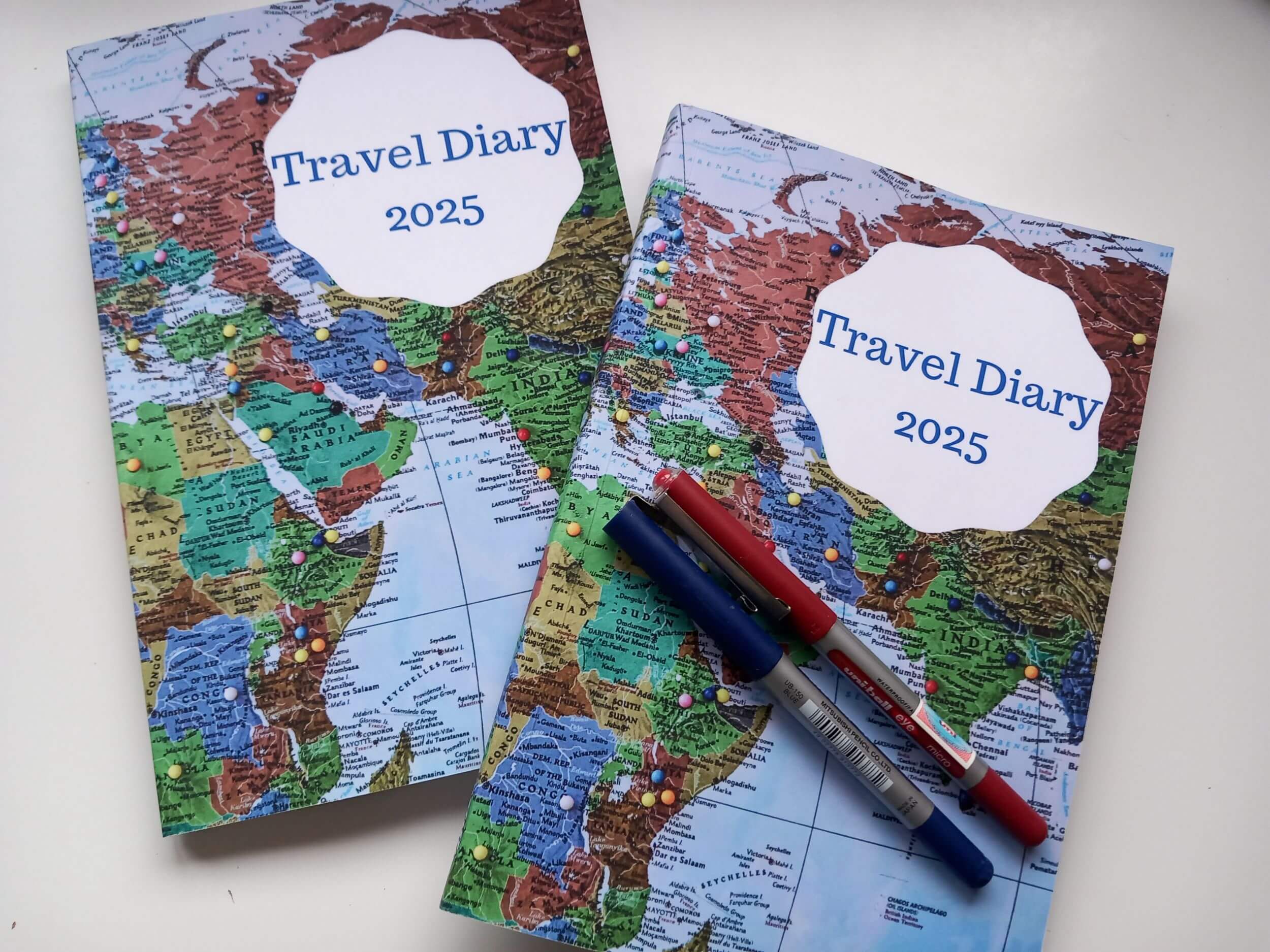 This photo shows two copies of the 2025 travel diary with two pens lying diagonally across one of them