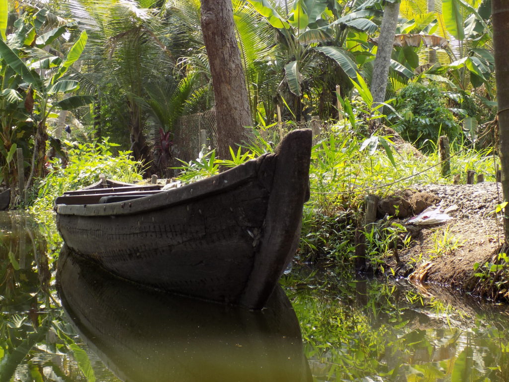 This photo shows a black boat moored in a backwater. The water is very still and the boat is reflected perfectly in the water