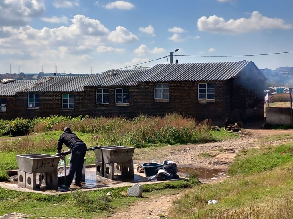 This image shows a row of dormitory accommodation with a man in the foreground drawing water from a well