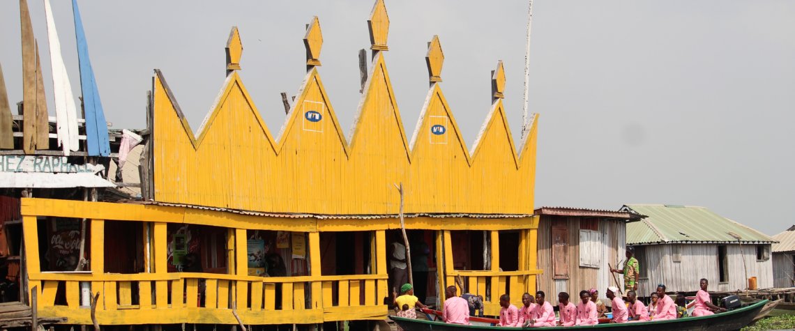 This photo shows a boat full of people dressed in pink arriving at a bright yellow-painted restaurant