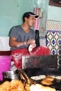 This photo shows a chef sitting on a tall stool shaping doughand frying doughnuts in a huge pan of hot oil