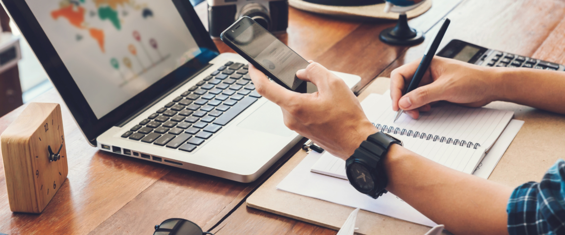 This photo shows someone holding a phone and looking at their laptop while making notes